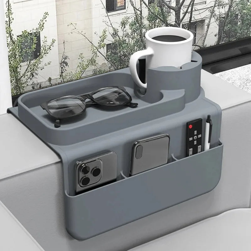 A gray silicone sofa armrest tray with a white mug, sunglasses, and side pockets holding a phone and remote.